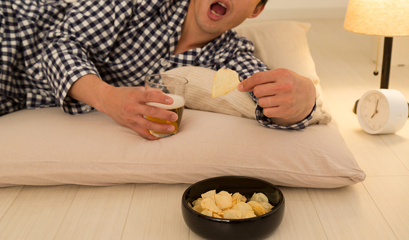 寝ながらポテトチップスとビールを食す男性