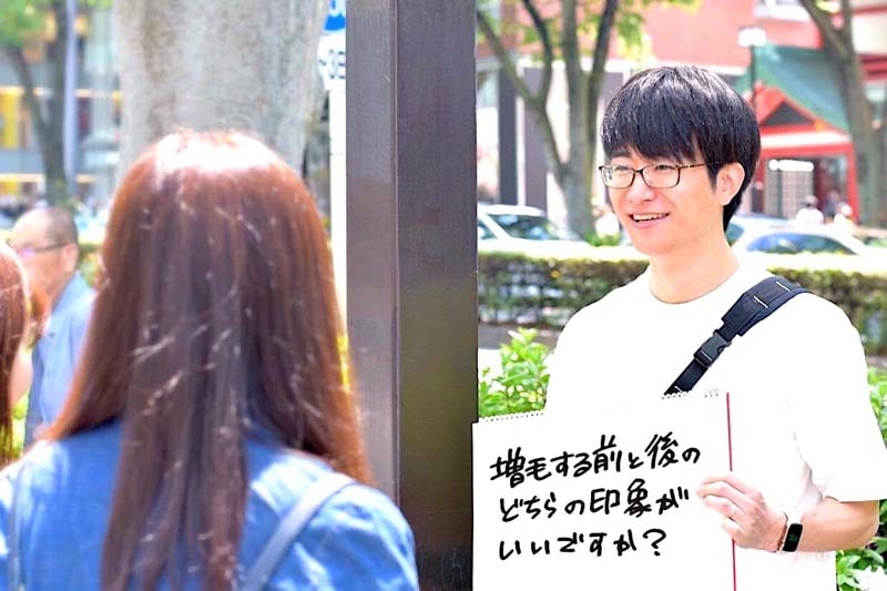 「増毛する前と後、どちらの印象がいいですか？」というフリップを持って女性に質問しているコクブさんの姿