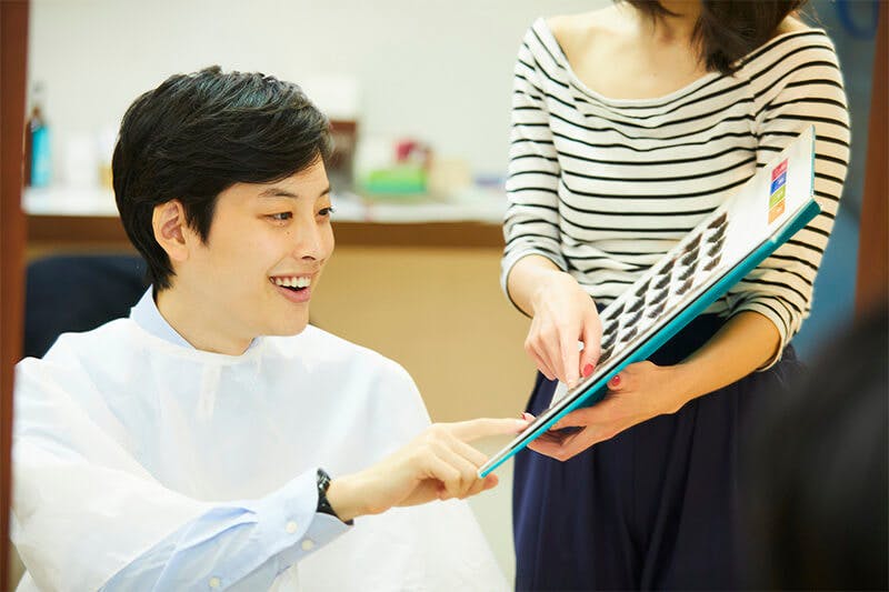 美容師にリストを見せられ、好みのヘアスタイルを笑顔で選ぶ男性